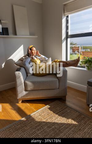 Asian woman reclining on gray armchair at home near window, wearing headphones and mustard dress. Sofa, sunlit, relaxation, cozy, minimalist, greenery, art Stock Photo