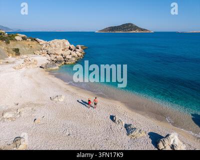 Gentle waves lap against the pristine shores of Gerenlik Beach in Kas, Antalya, where two people stroll along the white pebbled beach under a clear blue sky while the distant island beckons. Stock Photo