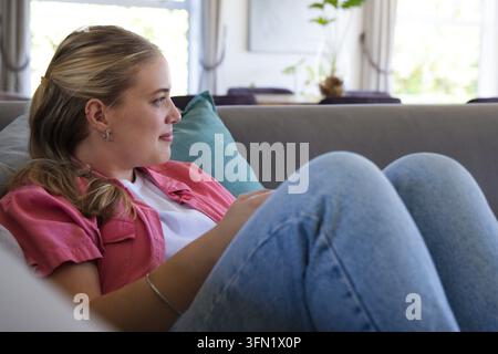 Adult woman reclining on grey sofa leaning against teal cushion holding smartphone at home Stock Photo