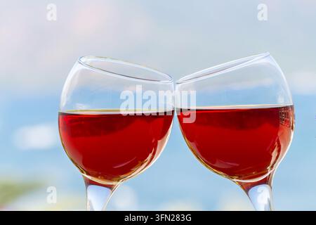 Close view of two stemmed glasses of rose wine leaning together in a toast with a softly blurred blue background suggesting a relaxed outdoor setting Stock Photo