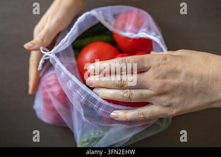 Top view of a hand resting on fresh red tomatoes and peppers inside a reusable mesh produce bag on a wooden table. Concept of sustainable grocery shop Stock Photo