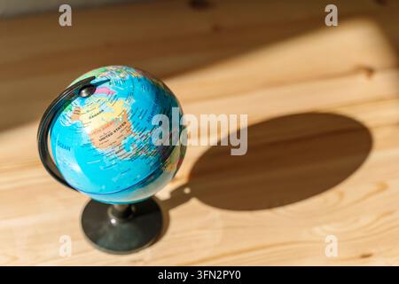 Globe on a wooden surface representing geography and education. Globe of Earth on a black stand placed on a light wooden surface against a textured white wall, illuminated by soft sunlight casting a shadow. Stock Photo