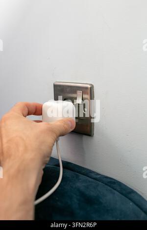 Close-up view of a person’s hand inserting a compact white electrical adapter or smart plug into a metal wall socket. Stock Photo