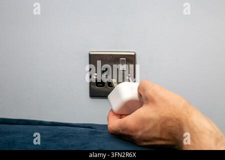 Close-up view of a person’s hand inserting a compact white electrical adapter or smart plug into a metal wall socket. Stock Photo