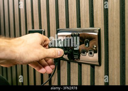 Close-up view of a person plugging a black UK three-pin electrical plug into a polished chrome double switched wall socket mounted on vertical panels Stock Photo