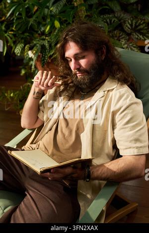 Thoughtful Man Writing in Notebook Sitting in Greenhouse Stock Photo
