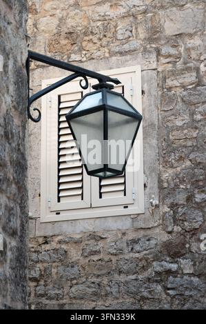 Weathered iron street lantern mounted beside shuttered window on textured limestone wall creating rustic historic Mediterranean exterior facade in Stock Photo
