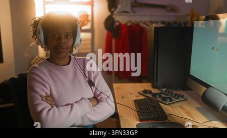 Young african american woman wearing white headphones typing on keyboard at wooden desk with monitor and audio mixer in studio; focus remote work. Stock Photo