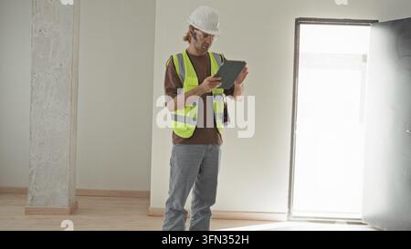 Man in white hardhat and high visibility vest taps tablet and looks up inspecting concrete column in building; focused inspection planning. Stock Photo