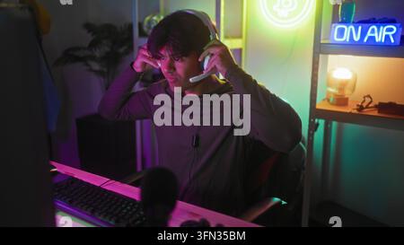 Man adjusts white headset and types on keyboard at a computer desk in studio with neon on air sign, gaming chair and shelf lighting; concentration. Stock Photo