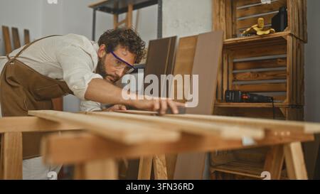 Man carpenter wearing apron and goggles sands wooden plank with hand in studio; craftsmanship concentration. Stock Photo