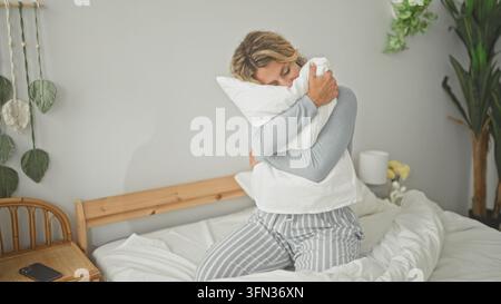 Woman hugging pillow, hands clasped, kneeling on bed in a bright bedroom with plants, nightstand and lamp; serenity. Stock Photo