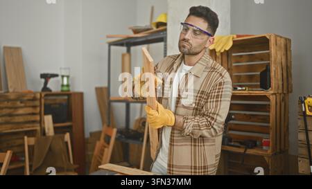 Man carpenter examining a wooden plank with gloved hands and safety goggles in a building workshop; concentration craftsmanship. Stock Photo