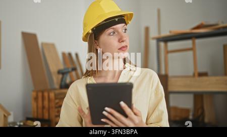 Woman carpenter wearing a yellow hardhat holds a tablet and looks to the side inside a building; concentration planning. Stock Photo