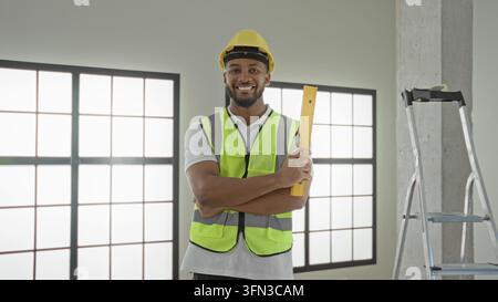 Man builder in hardhat smiles and holds a yellow measuring level with arms crossed revealing forearm inside a building; confidence professionalism. Stock Photo