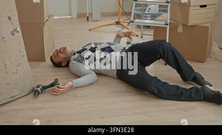 Man lying unconscious on floor with hand near cordless drill beside stacked cardboard boxes and ladder in building interior new home moving scene; acc Stock Photo