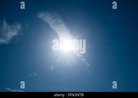 A bright sun flare shines behind a thin wispy cirrus cloud in a deep blue sky, shot from a low angle, with a faint aircraft contrail and small scatter Stock Photo