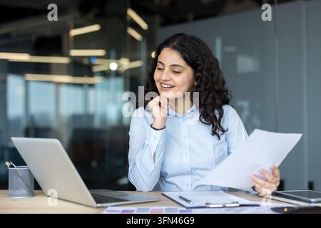 Young businesswoman smiling while reading a document at her office desk, working on a laptop with confidence and focusing on financial success and business growth Stock Photo