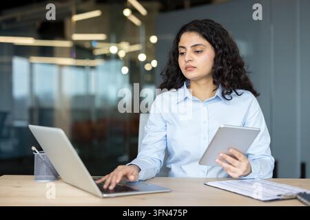 Young businesswoman working efficiently in a modern office. Simultaneously using a laptop and a digital tablet. Demonstrating productivity and technological integration in a busy professional setting Stock Photo