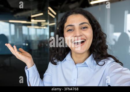 Young businesswoman with curly hair smiling broadly, gesturing with an open hand, and looking directly at the camera while making a video call or recording a vlog from a modern office environment Stock Photo