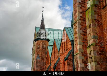 Close-up view of Ribe Cathedral featuring ornate red brick architecture, oxidized copper roofing, and a spire with cross against cloudy sky. Stock Photo