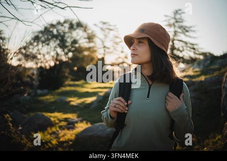 View of a woman trekking in the mountains wearing a bucket hat and backpack during golden hour in Tandil, Buenos Aires, Argentina. Stock Photo