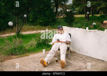 A man in a white T-shirt and trousers, dark baseball cap and sunglasses reclines in a folding chair beside a low white wall outdoors. He holds a phone Stock Photo