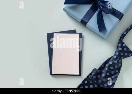 Blank white greeting card with blue envelopes, wrapped gift and patterned necktie arranged on a light blue background. Father's Day message mockup and Stock Photo