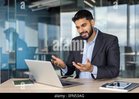 Young indian businessman discussing project online, using a laptop for a virtual meeting or conference, working from a modern corporate office interior Stock Photo