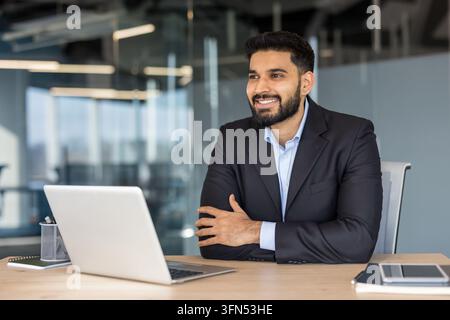 Positive young indian businessman sitting at an office desk with a laptop, confidently looking away and smiling, embodying professionalism and success in a modern corporate environment Stock Photo