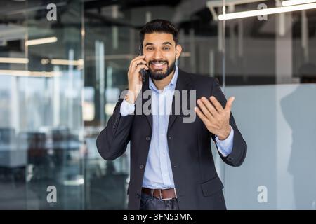 Young indian businessman smiling and talking on his smartphone, discussing business while standing in a contemporary glass office building, actively communicating and connecting Stock Photo