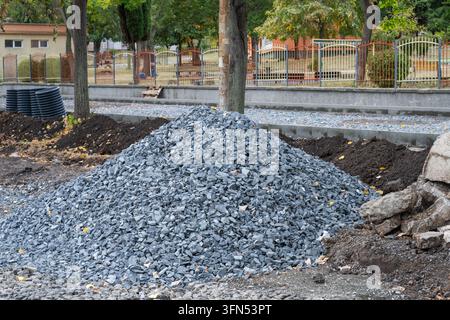 A large conical pile of blue-grey crushed stone aggregate sits on a dirt roadbed beside fresh soil and concrete curbs during an urban park sidewalk re Stock Photo