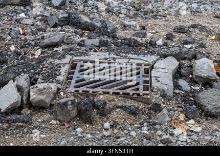 A rectangular cast iron storm drain grate sits surrounded by crumbling concrete kerbing, crushed gravel, and broken asphalt debris during urban road r Stock Photo