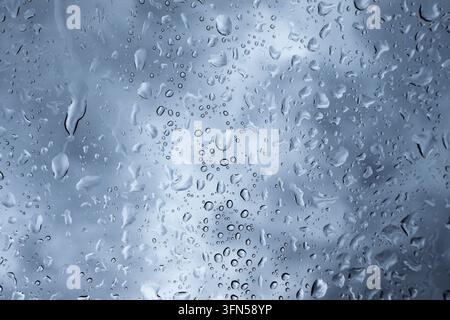 Detailed macro shot of raindrops streaming and clinging to a glass pane against an overcast blue sky Stock Photo