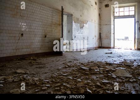 The interior of a dilapidated and abandoned room features walls covered in old, largely broken white tiles. The floor is entirely littered with debris Stock Photo