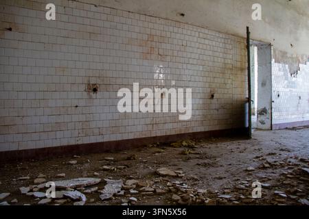A side view shows a long wall covered with old, dirty square white tiles. The concrete floor is scattered with rubble and shards, while an open doorwa Stock Photo