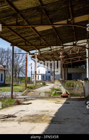 A perspective shot shows a long canopy made of rusty iron covering a concrete platform. In the background, a white two-story building is visible, whil Stock Photo