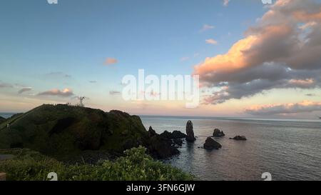 The green Seopjikoji headland and its lighthouse overlook dramatic basalt sea stacks in the calm sea. Soft pink clouds fill the golden-hour sky near S Stock Photo