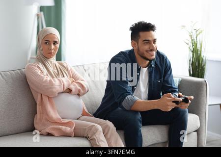 Lack Of Care. Upset Expectant Muslim Woman Sitting On Couch, Feeling Offended To Her Husband That Playing Video Games At Home, Indifferent Future Fath Stock Photo