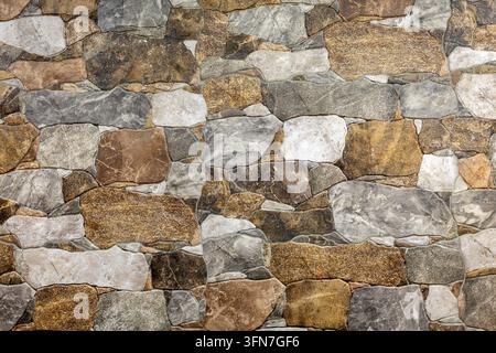 Natural stone wall made of irregular gray, brown and beige rocks, rustic masonry surface with varied shapes and detailed mineral texture. Stock Photo