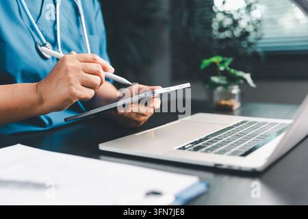 Medical professional using a tablet stylus beside a laptop and stethoscope, illustrating electronic health records, digital documentation, clinical wo Stock Photo