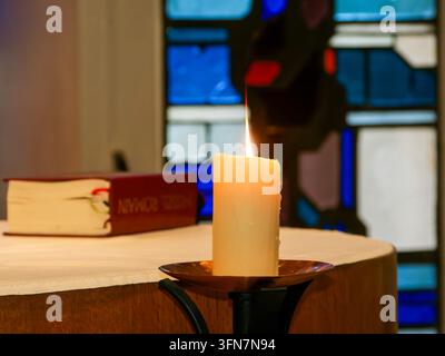 Lit white candle on holder beside a religious book on an altar with stained glass background. Stock Photo