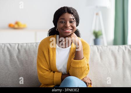 Domestic lifestyle concept. Relaxed young black lady in casual outfit sitting on couch in living room, smiling at camera, copy space. Happy african am Stock Photo