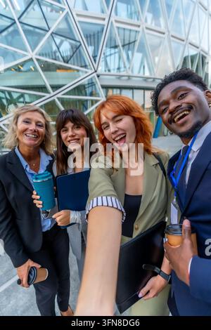 Diverse business professionals smiling and having fun taking a selfie outside a modern office building, celebrating teamwork and success during a brea Stock Photo