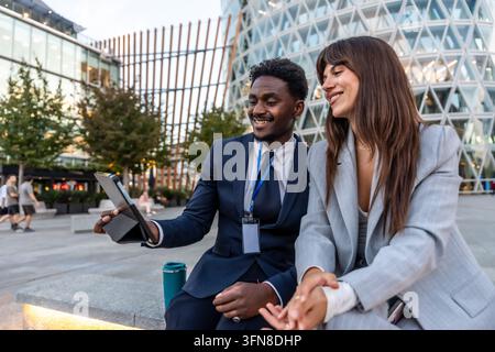 Two smiling diverse business professionals engaging in a discussion while looking at a digital tablet, sitting on a bench in a modern urban environmen Stock Photo