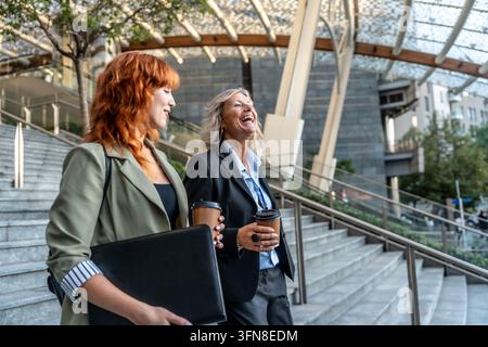 Two professional business women, a young one and a senior one, walking up city stairs while holding takeaway coffee cups, engaging in cheerful convers Stock Photo