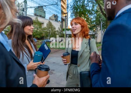 Diverse business professionals standing outdoors, engaging in conversation and discussing work topics during a break, holding coffee cups in an urban Stock Photo