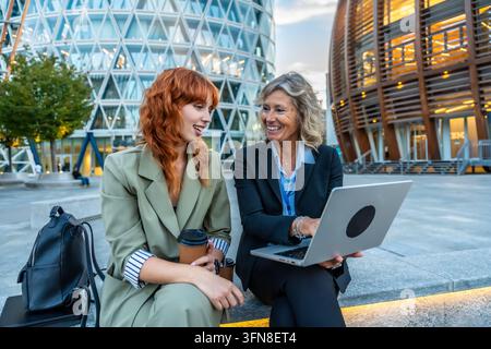 Two smiling businesswomen interacting while working on a laptop outdoors, discussing business strategy with coffee cups and a backpack in a contempora Stock Photo