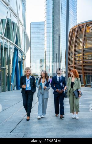 Diverse business professionals walking and discussing in an urban financial district, holding coffee cups and folders, showing collaboration and teamw Stock Photo
