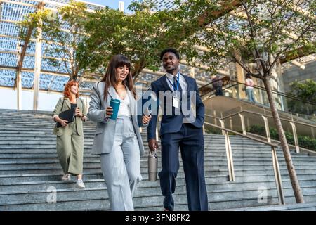 Three diverse business professionals walking down urban stairs, chatting and smiling while holding documents and reusable coffee cups, symbolizing tea Stock Photo